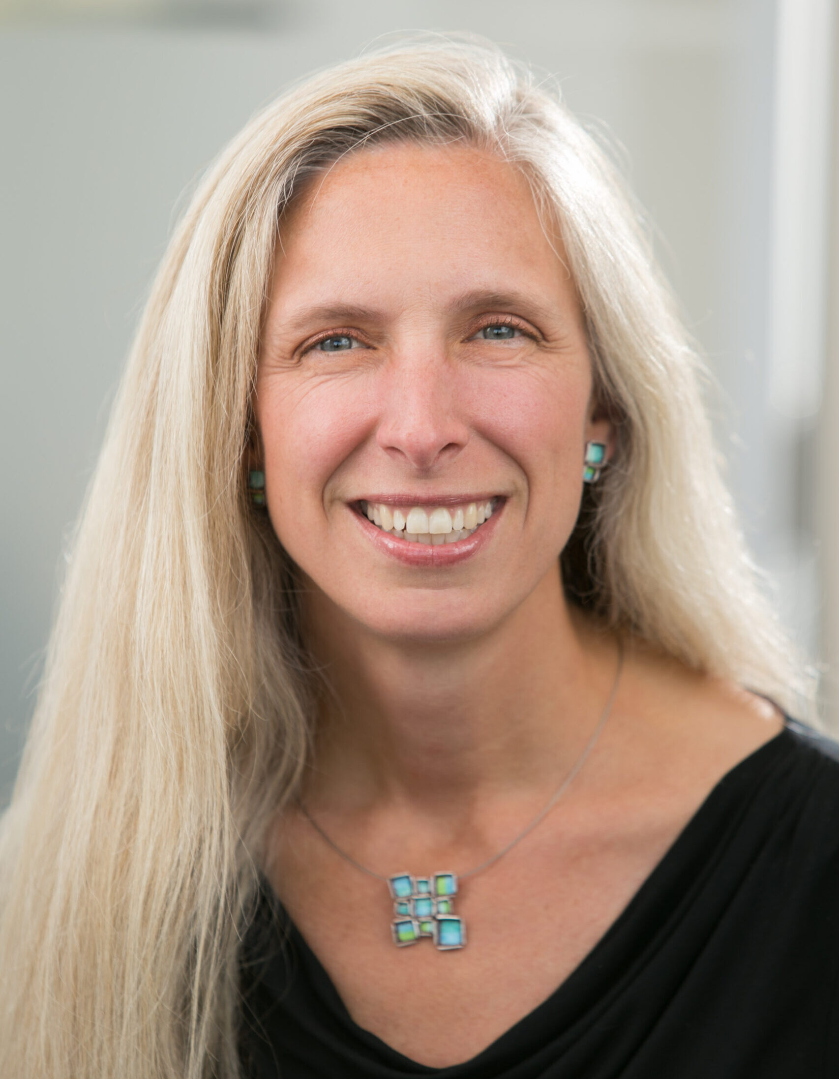 smiling woman with long blonde hair wearing black top and turquoise pendant necklace, smiling