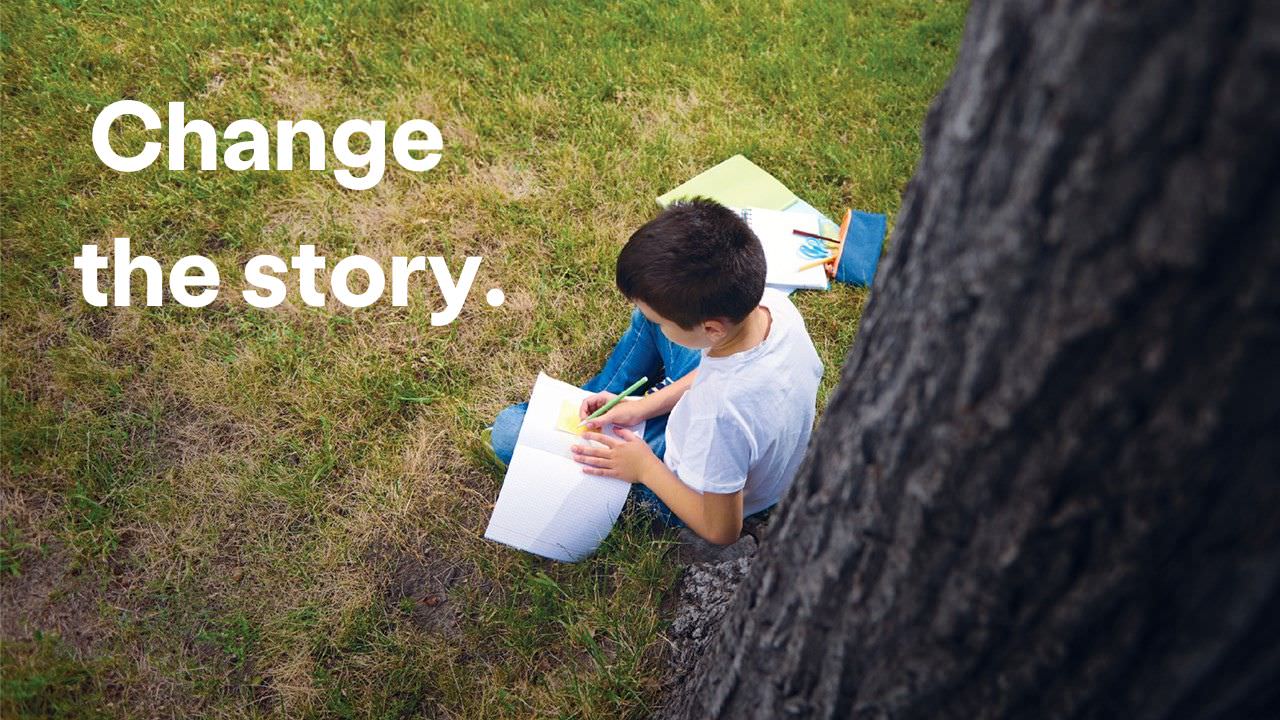 Boy in white shirt sitting on grass under a tree writes a story. Text reads Change the Story.