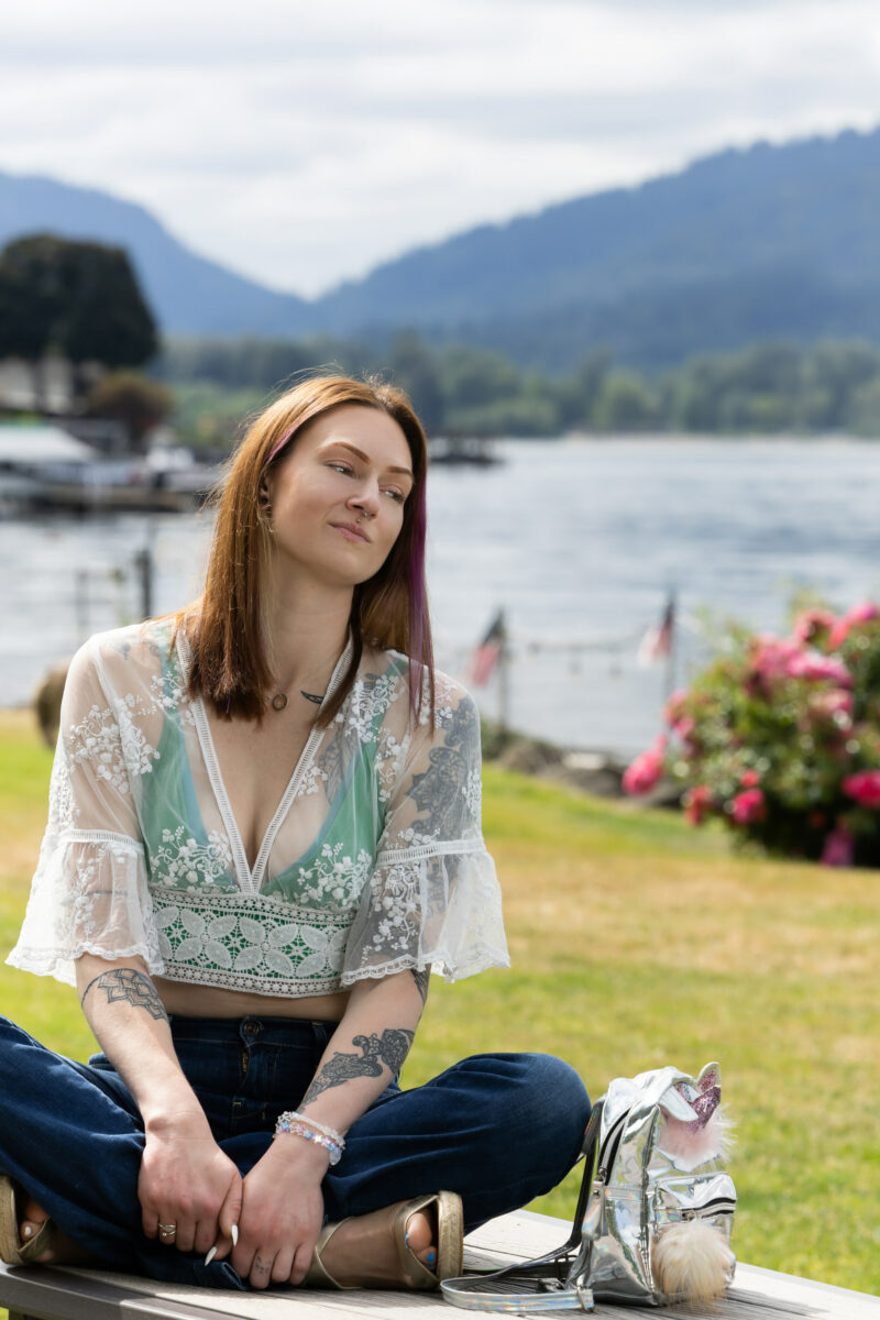 Un membro di Empowered Voices indossa un top di pizzo e verde e jeans blu, sull'erba con il lago e le montagne sullo sfondo