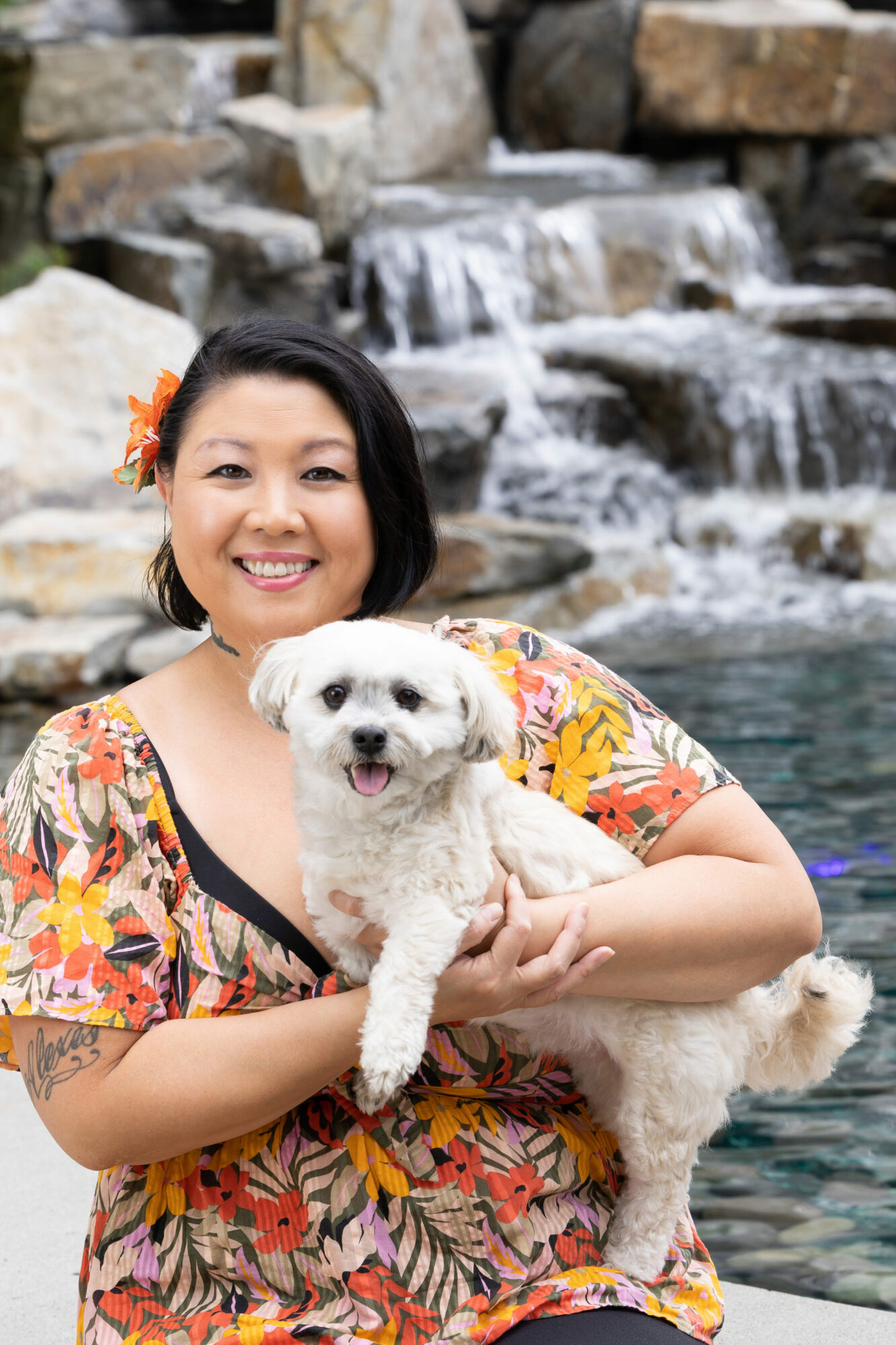 Empowered Voices-Mitglied trägt eine bunte Blumenbluse und hält einen kleinen weißen Hund mit einem mehrstufigen Felsenwasserfall im Hintergrund