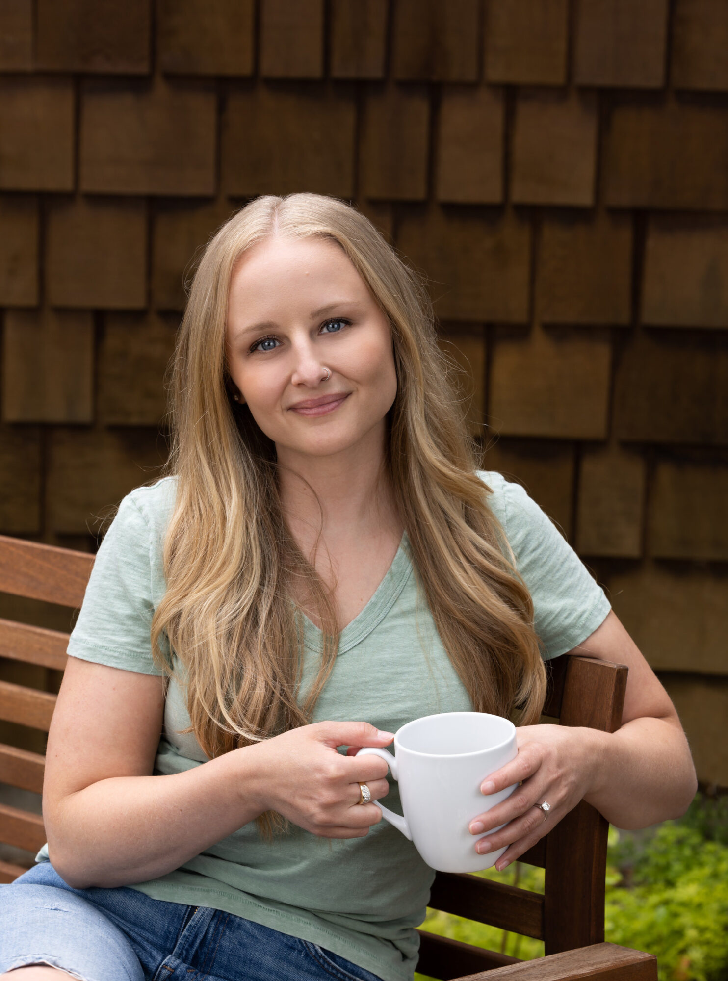 Empowered Voices member wearing a light green v-neck t-shirt sitting on a brown wooden slatted bench, holding a white coffee cup with dark brown cedar shake wall in background.
