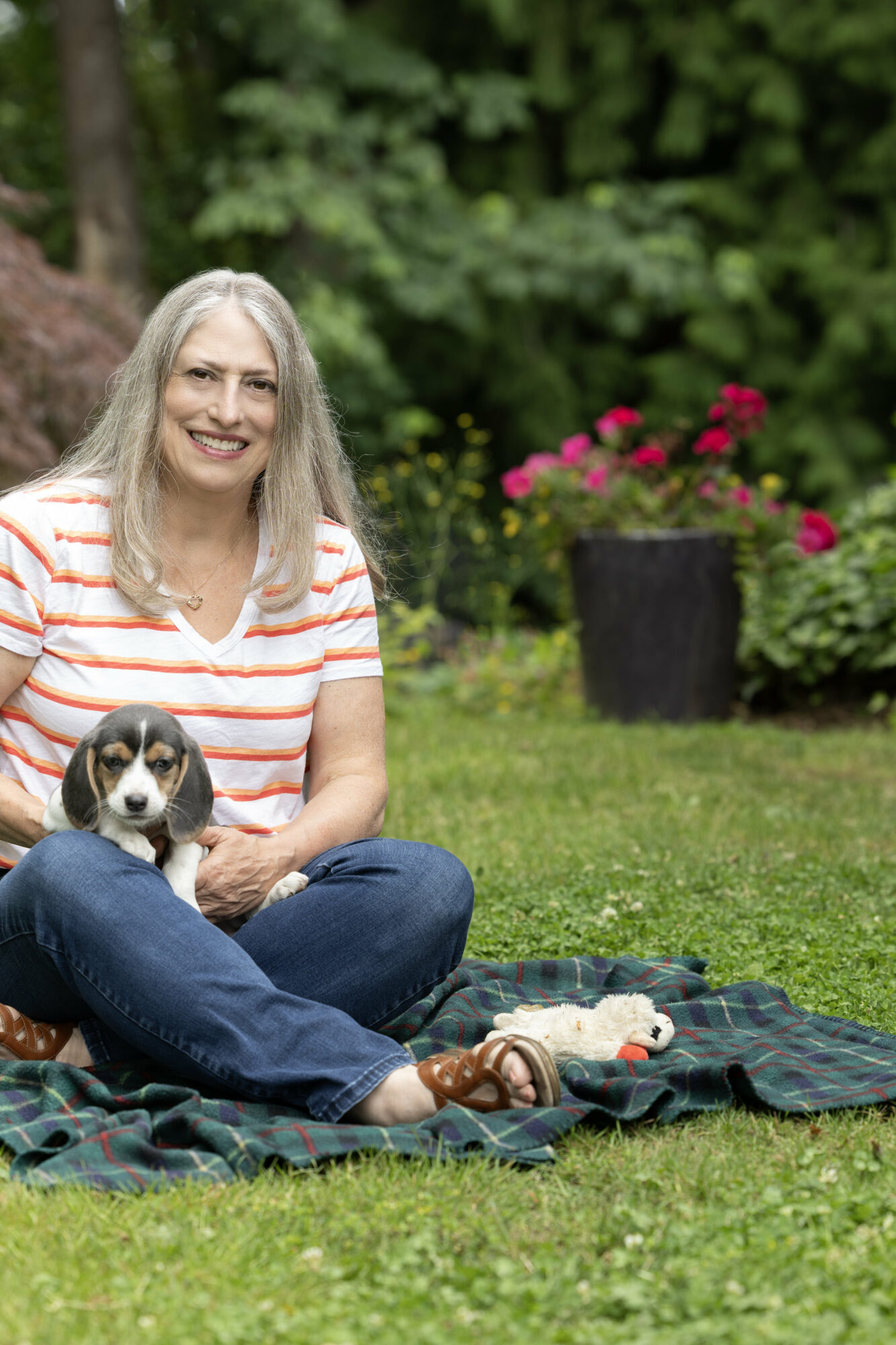 Empowered Voices member wearing a orange and white striped v-neck t-shirt and blue jeans with beagle puppy on lap on a dark green plaid blanket on grass with pink potted begonias and forested green background