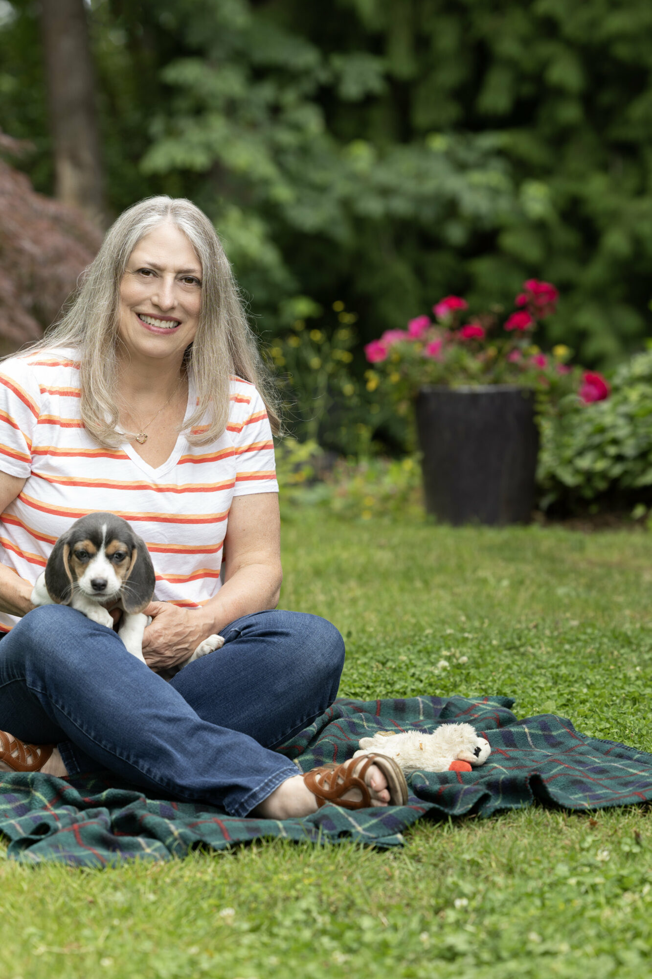 Membro do Empowered Voices vestindo uma camiseta listrada laranja e branca com decote em V e jeans azul com cachorrinho beagle no colo em um cobertor xadrez verde escuro na grama com begônias em vasos rosa e fundo verde florestado