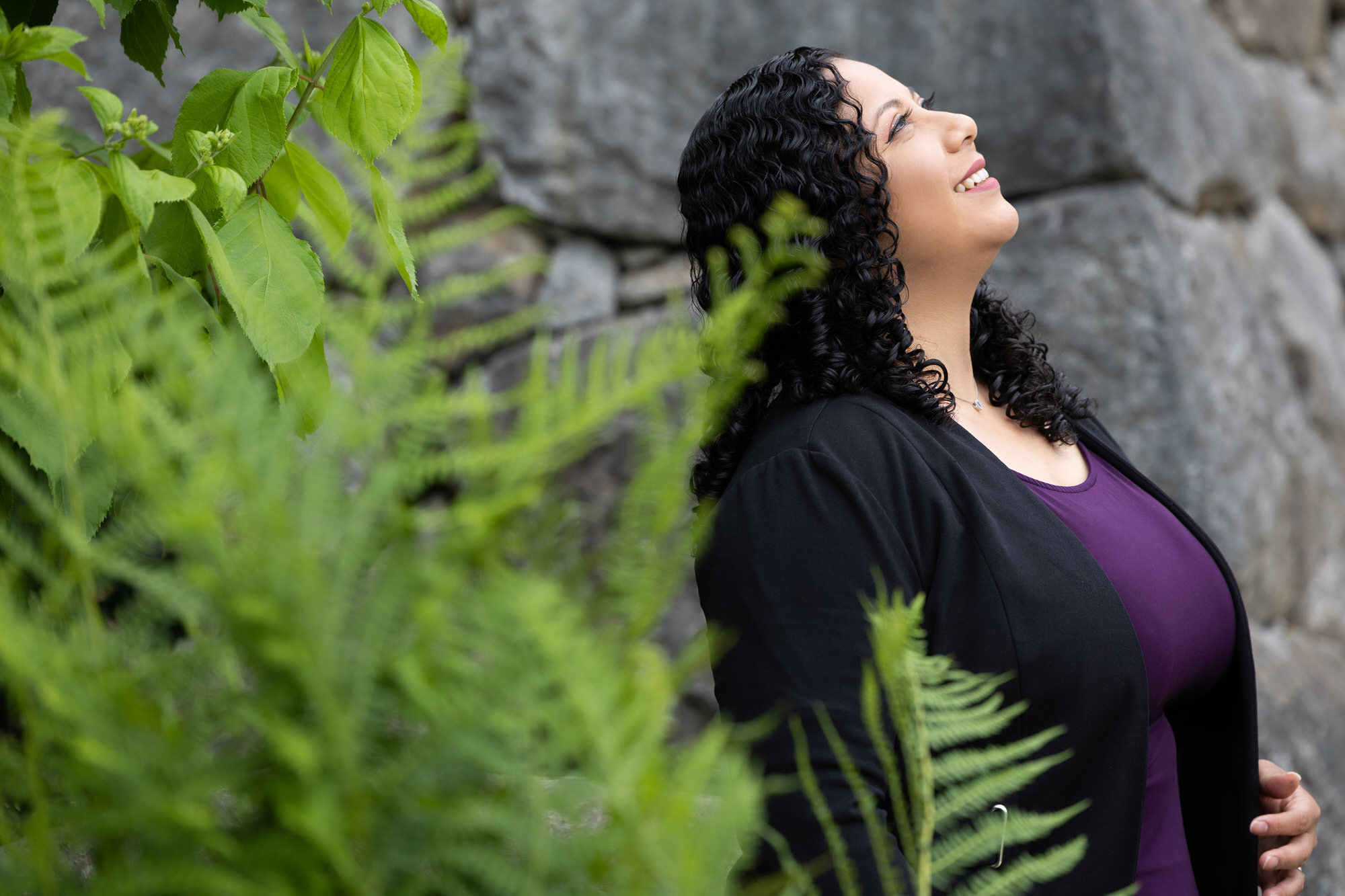 Un miembro de Empowered Voices que viste una blusa de color púrpura real y un cárdigan negro sonríe y mira hacia el cielo, apoyado contra una pared de piedra gris cerca de un follaje de helechos verdes.