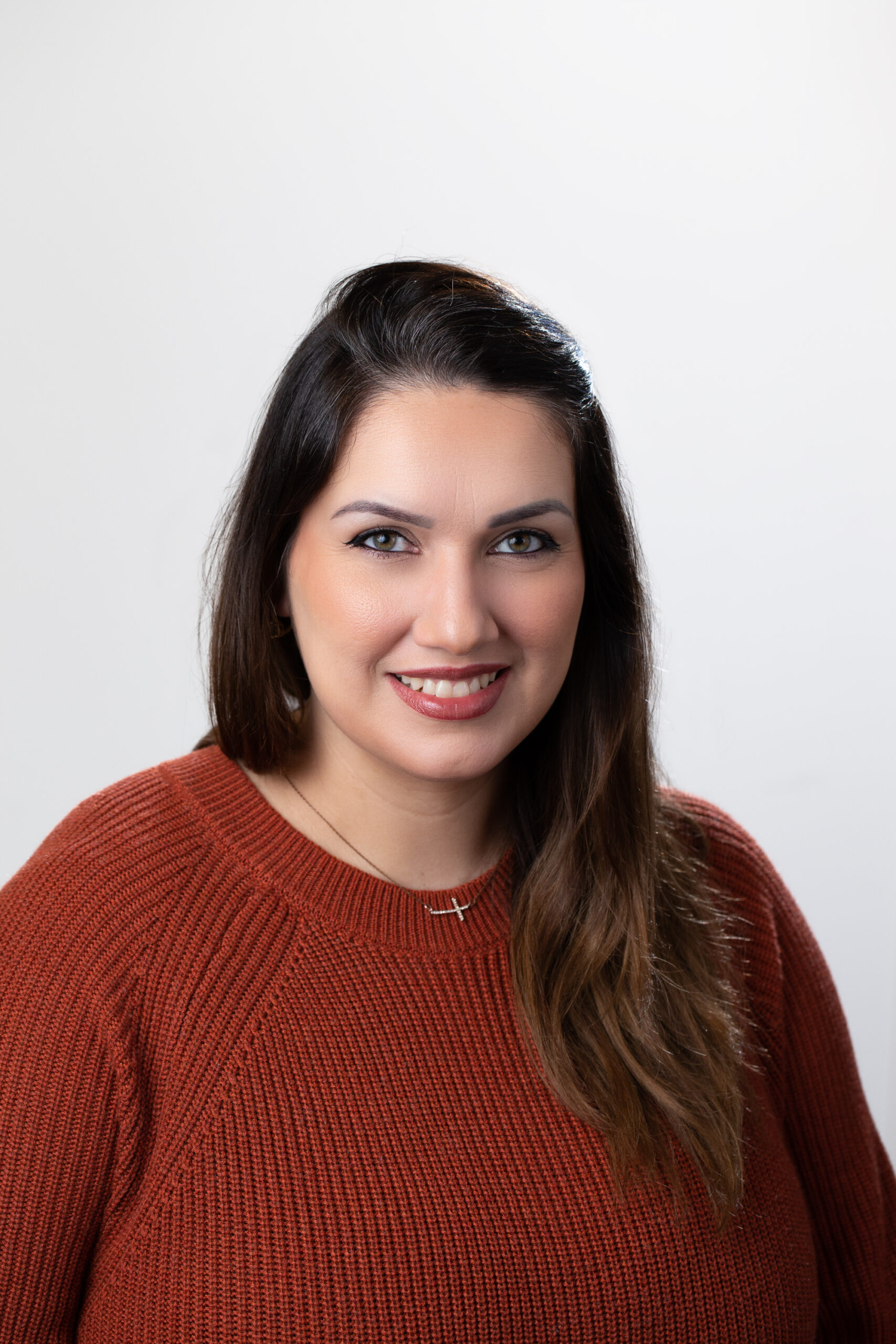 Une femme souriante aux longs cheveux noirs porte un pull orange et un collier doré.