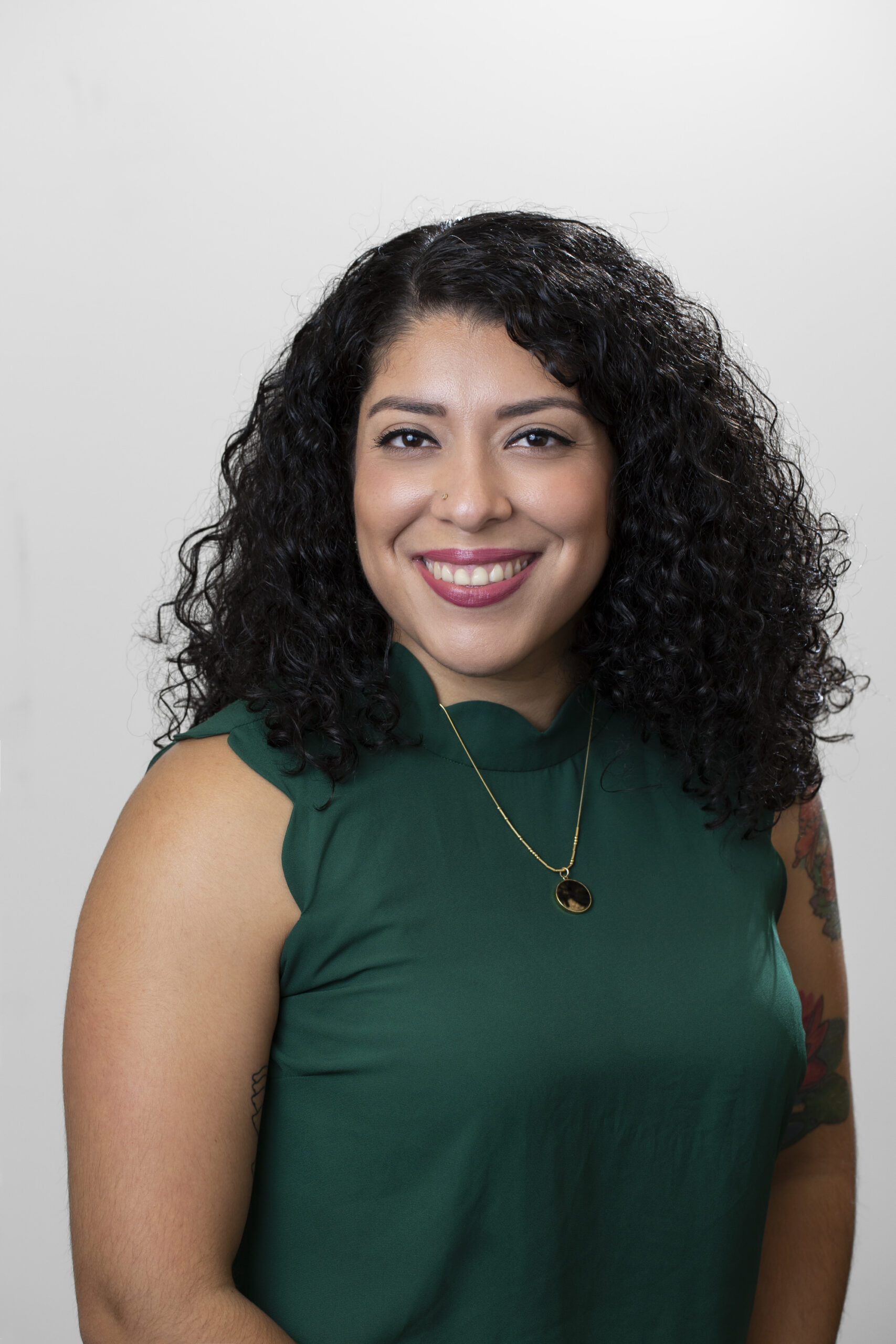 Femme souriante aux longs cheveux noirs bouclés portant un chemisier vert sans manches et un collier en or avec pendentif noir