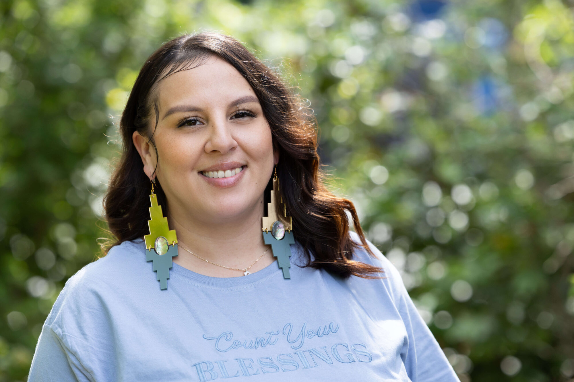 smiling woman with long dark hair, long blue and green earrings and wearing a light blue shirt