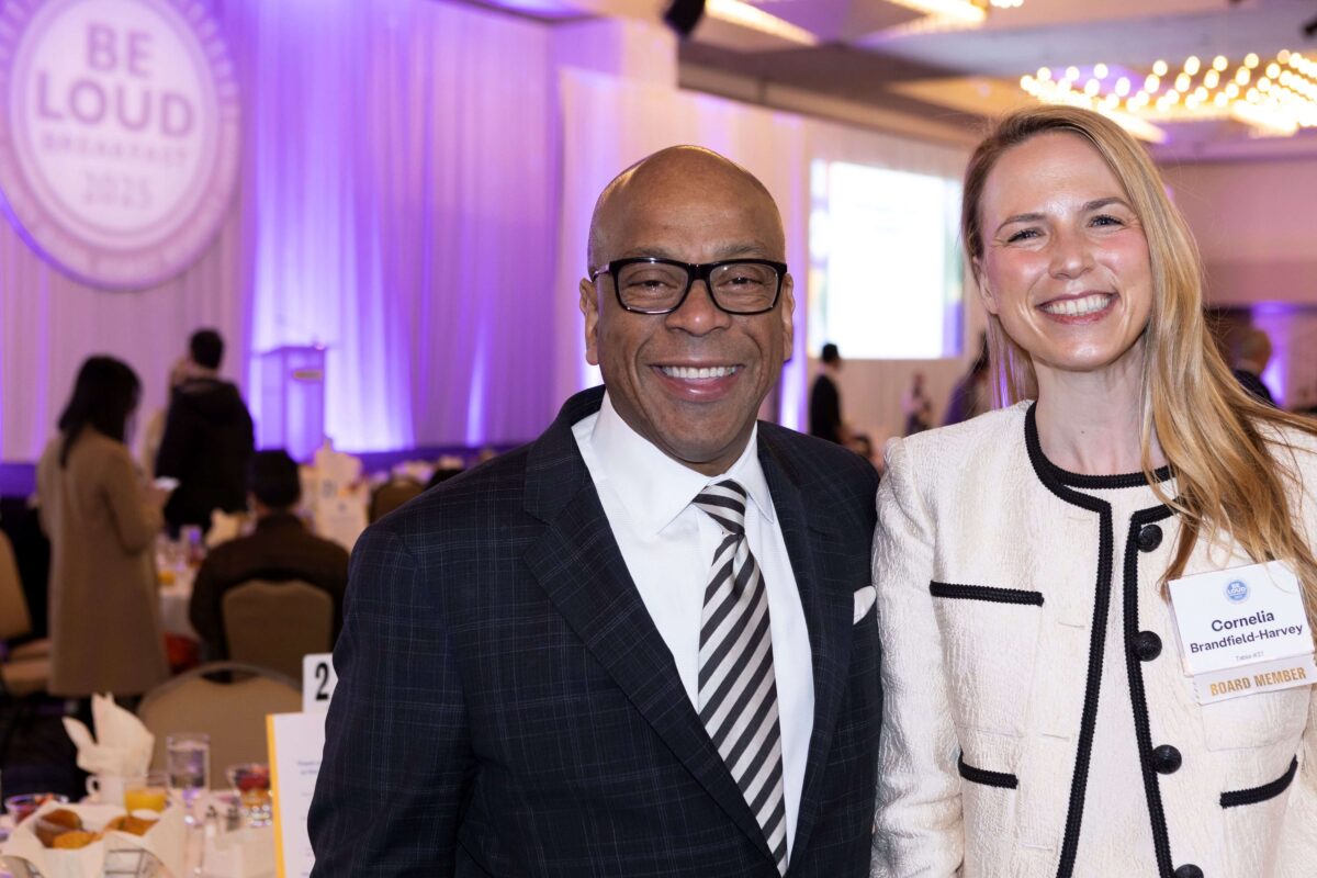 Smiling man in dark suit, black and white striped tie and white shirt and smiling woman with blonde hair, white jacket and top with black trim.