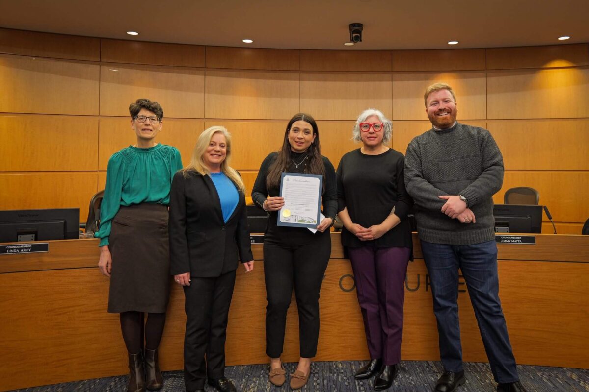 Group of five smiling people stand in front of a wood paneled wall and city council chamber podium. At center a woman with long dark hair holds a document with gold seal.