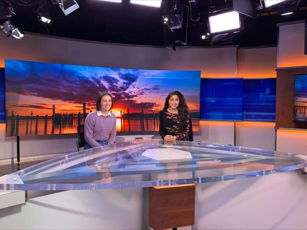 Two smiling women seated in television studio behind glass desk. Woman on left wears lavender sweater with white collar. Woman on right wears floral dress.