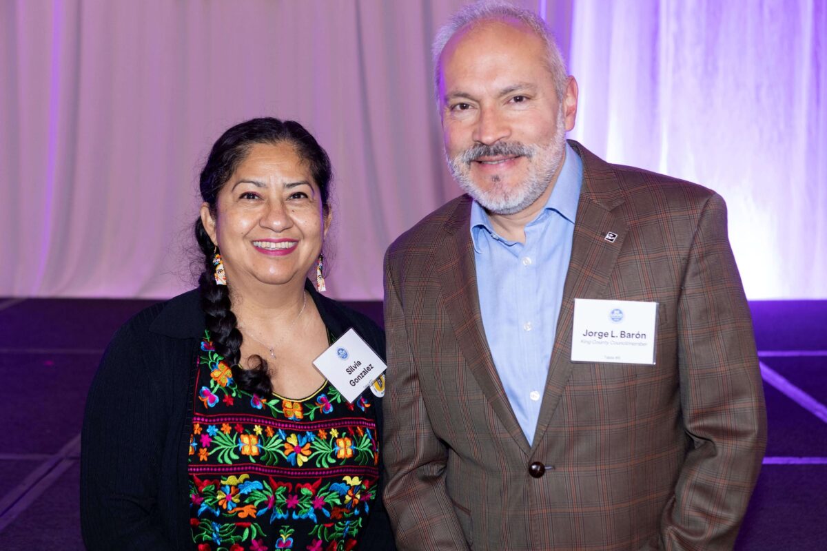 smiling woman with braided dark hair, long earrings wearing a bright embroidered top and black jacket and smiling man with gray hair, beard and mustache wearing a brown plaid jacket and light blue shirt