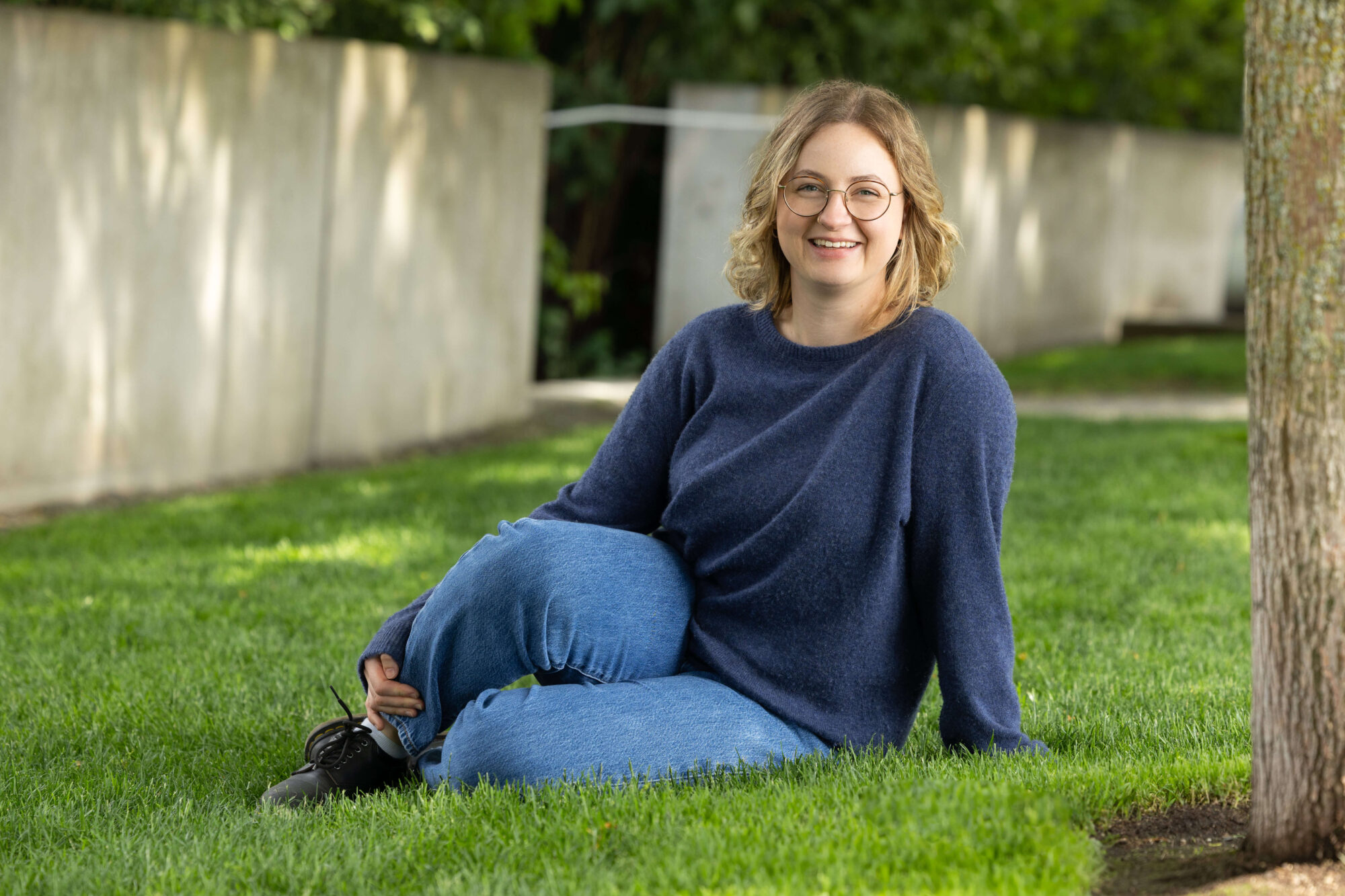smiling woman with medium length blonde hair, glasses wearing a blue sweater and jeans sitting on grass