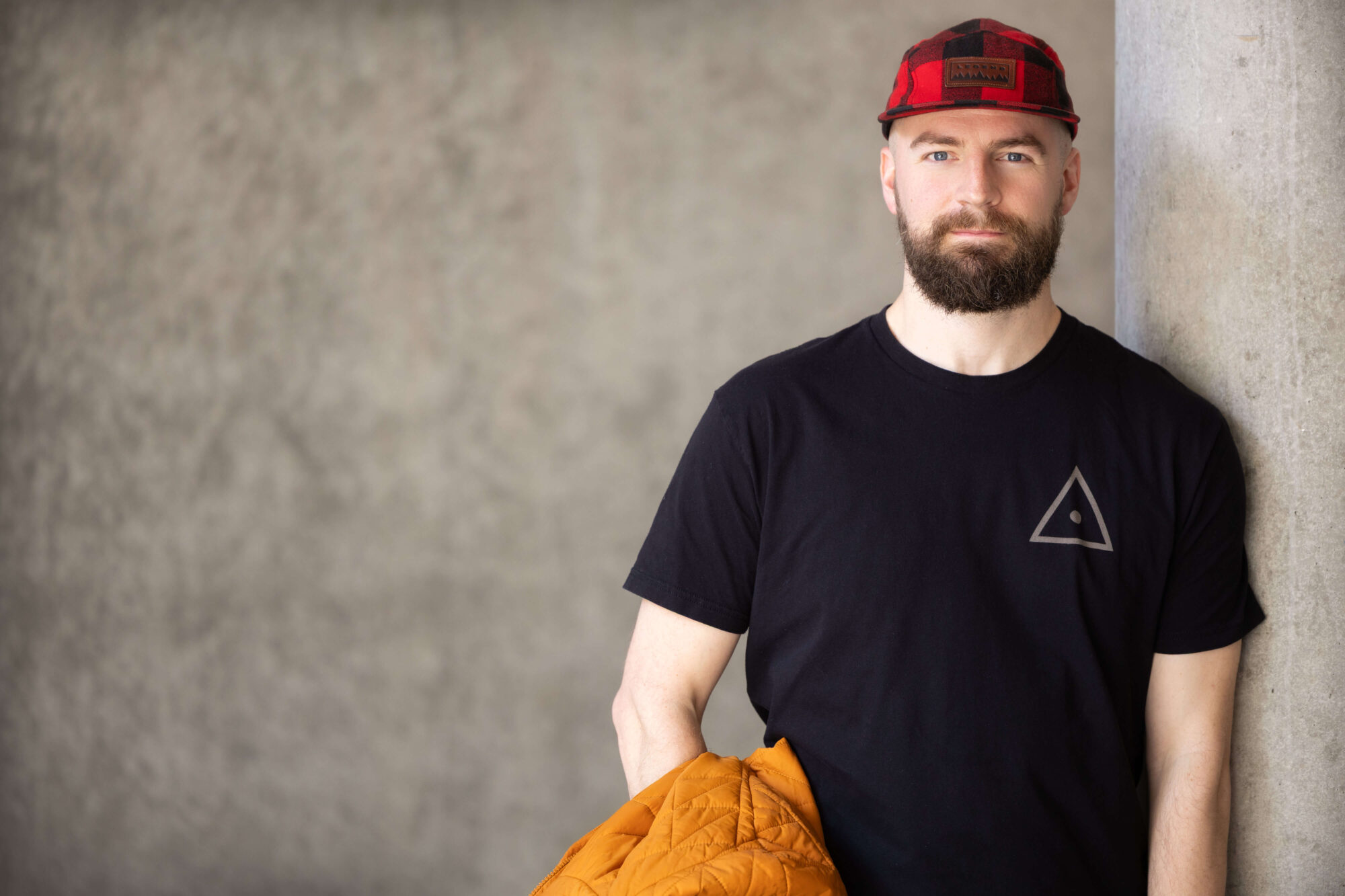 serious faced man with brown beard and mustache, wearing a red plaid cap and black t-shirt