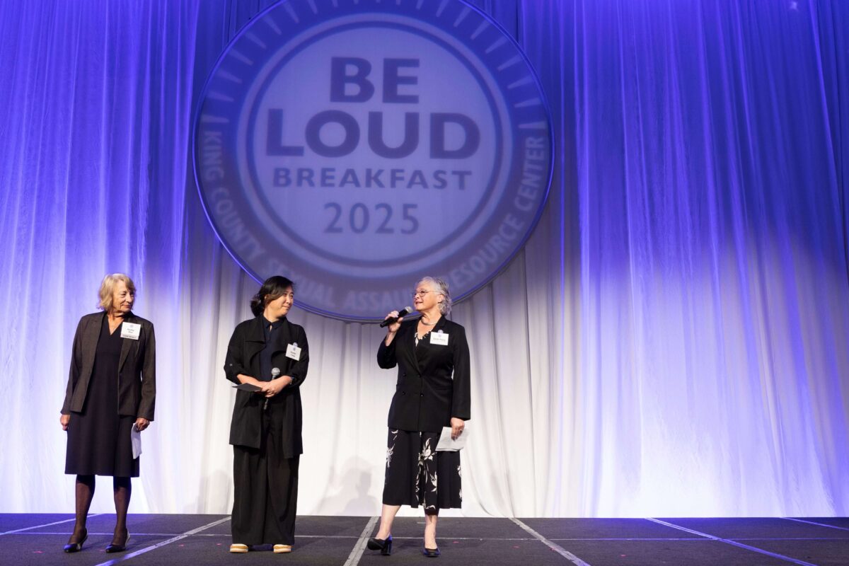Three women on a stage with purple backdrop saying BE LOUD Breakfast 2025 King County Sexual Assault Resource Center. All wear dark jackets, one holds microphone and is speaking while other two look her way.