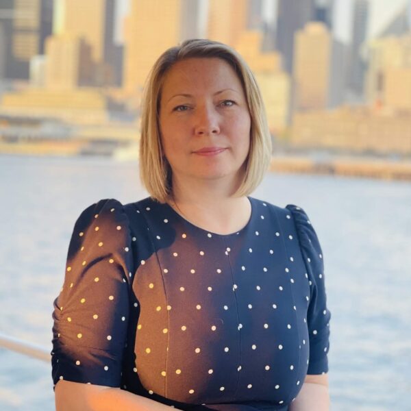 woman with light hair wearing a navy blue and white polka dot top against backdrop of water and city skyline