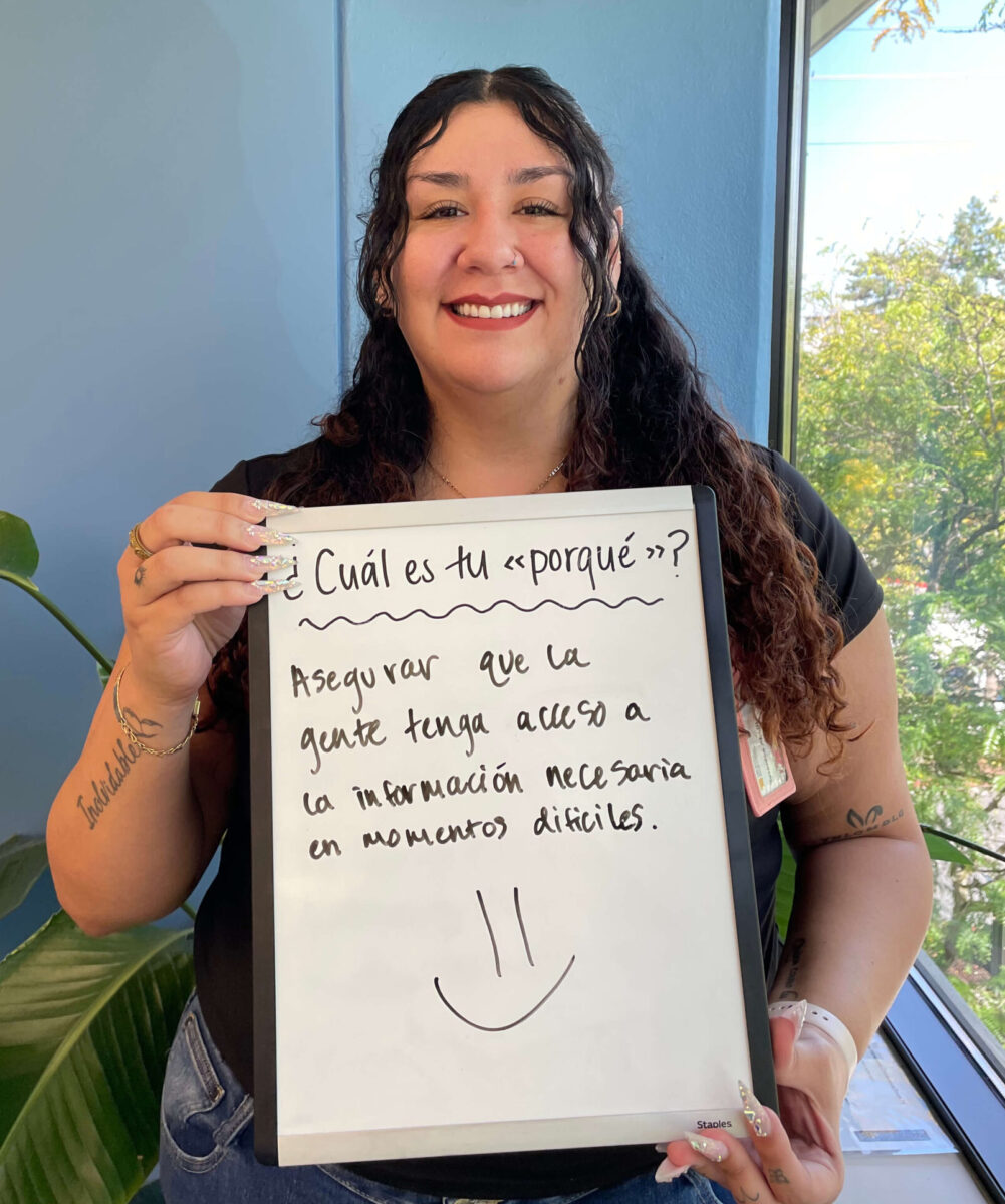 smiling woman with long curly hair holding a sign in Spanish reading“Asegurar que la gente tenga acceso a la información necesaria en momentos difíciles.” In English:“To ensure that people have access to the information they need during difficult times.”