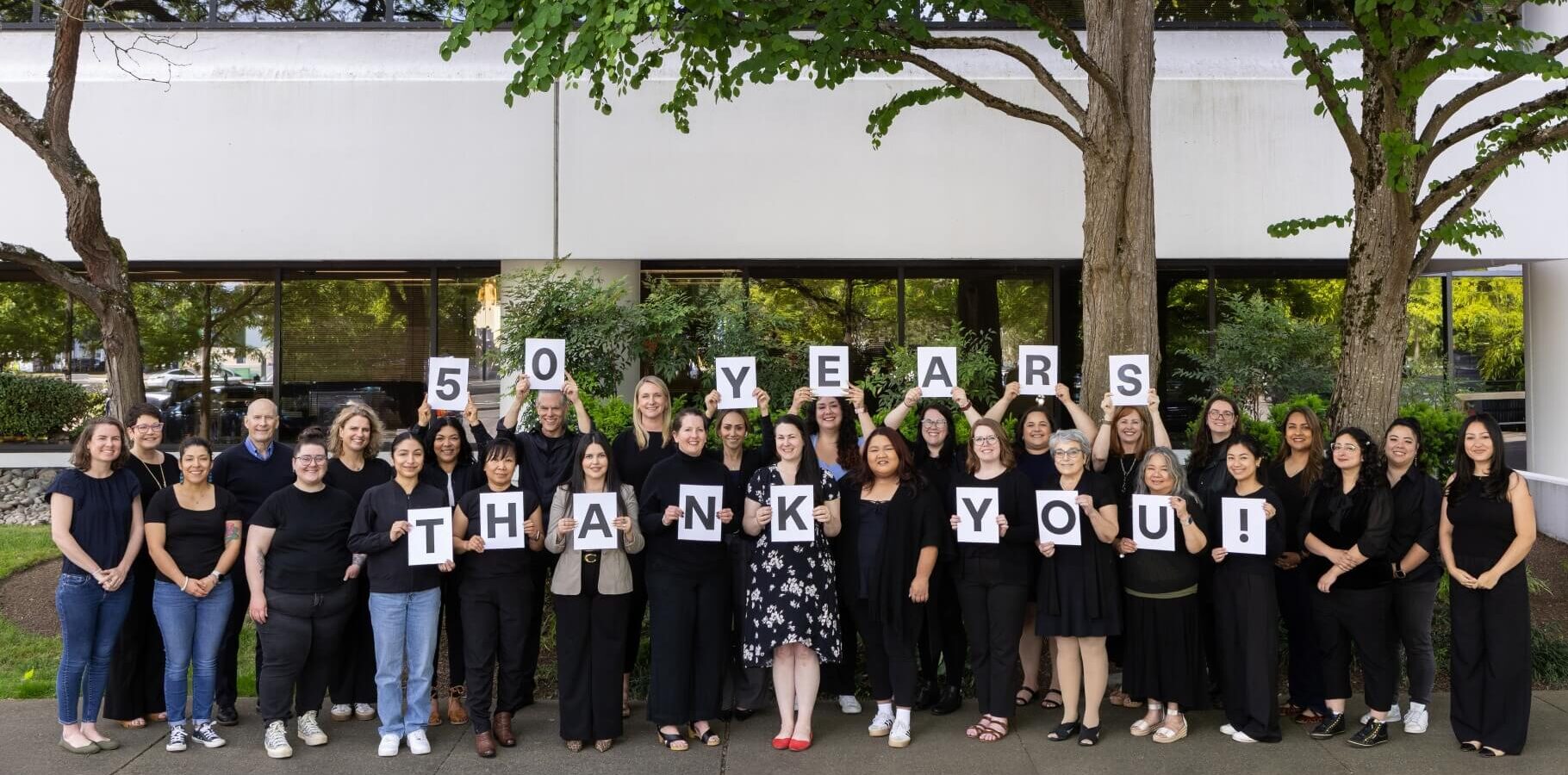 Group of people outdoors in front of office building holding individual letters that spell out the phrase "50 years thank you!"