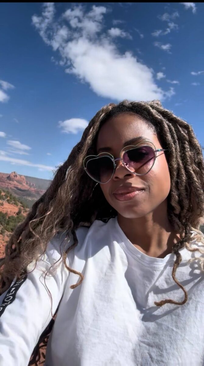 woman with brown skin, long hair, heart-shaped sunglasses and white shirt outdoors on a blue sky day