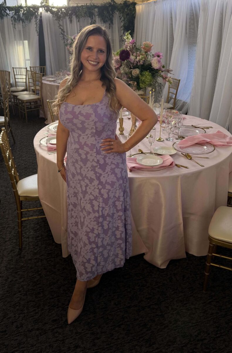 Une femme aux longs cheveux blonds, vêtue d'une robe lavande, se tient devant une table recouverte d'une nappe couleur crème.